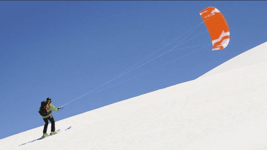 Person on skis kites up a snow-covered hill under a blue sky. Kite skiing in a winter landscape.