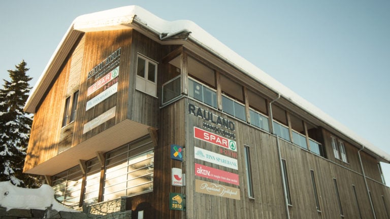 Träbygning med snø på taket, skilt for Rauland Handelshus, Spar og Tinn Sparebank, vinterdag.