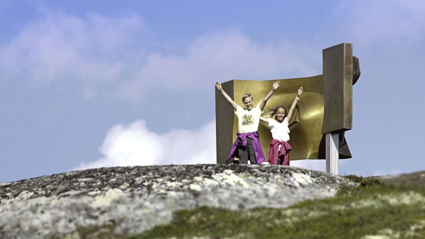 To barn foran en skulptur på fjellet med blå himmel i bakgrunnen.