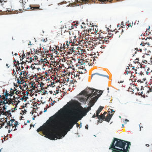 Mange mennesker samlet til skibegivenhet i snøen, med rampe og basseng, under en klar himmel.