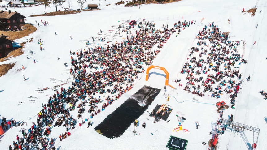 Mange mennesker samlet til skibegivenhet i snøen, med rampe og basseng, under en klar himmel.