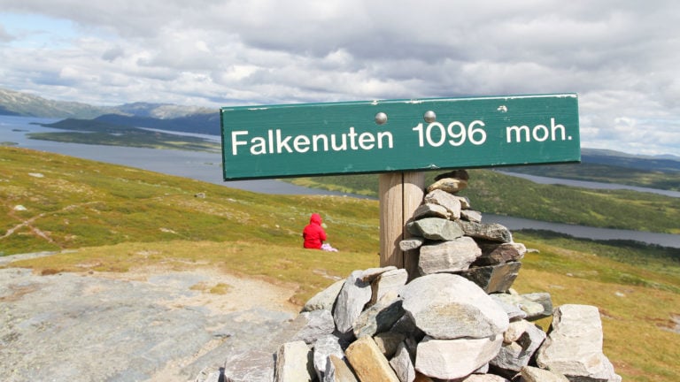 Fjelltopp med skilt Falkenuten 1096 moh. og vakker utsikt over innsjø og dal under skyet himmel.