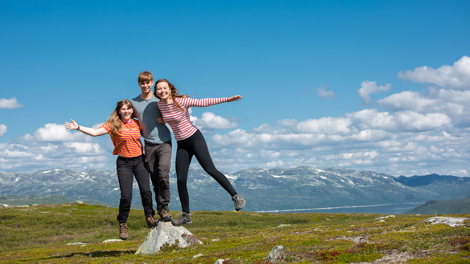 Gruppe på tre venner nyter solfylt tur med vakker utsikt over norske fjell.
