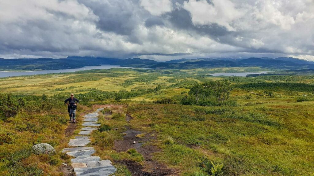 Vandrer på naturskjønn sti, med fjell og innsjø i bakgrunnen, under dramatiske skyer.