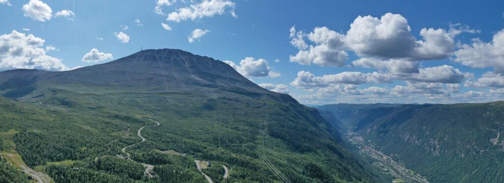 Panoramautsikt over Gaustatoppen med vakker blå himmel og dalen nedenfor dekket av frodig skog.