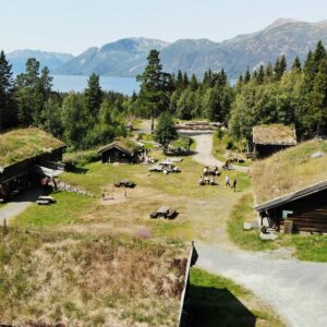Tradisjonell norsk gård med torvtak og fjordutsikt, omgitt av skog og fjell i naturskjønne omgivelser.