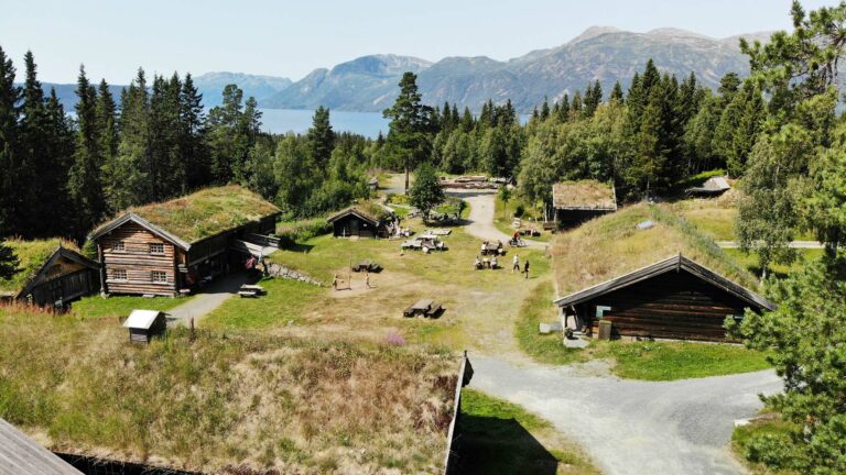 Tradisjonell norsk gård med torvtak og fjordutsikt, omgitt av skog og fjell i naturskjønne omgivelser.