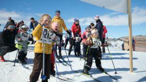 Barn på ski klare til start i et skirenn på snødekt bakke, omgitt av tilskuere under en blå himmel.