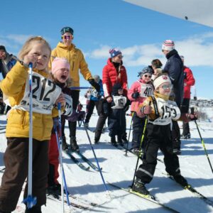 Barn på ski klare til start i et skirenn på snødekt bakke, omgitt av tilskuere under en blå himmel.