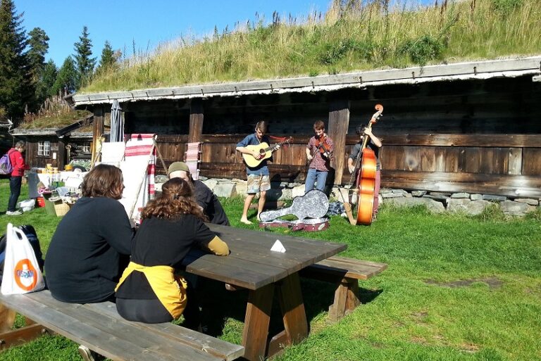 Utendørs sommerkonsert med tre musikere, gitar og kontrabass, mens folk ser på fra benk, norske hytter i bakgrunnen.