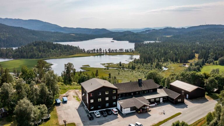 Hotel ved innsjø omgitt av skog og fjell i Norge, fredelig natur med parkerte biler foran bygningen.
