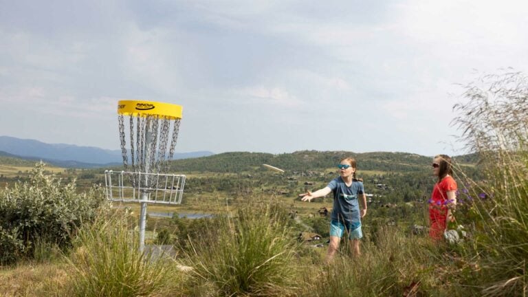 Barn spiller frisbeegolf i naturskjønne omgivelser, med kurv i bakgrunnen.