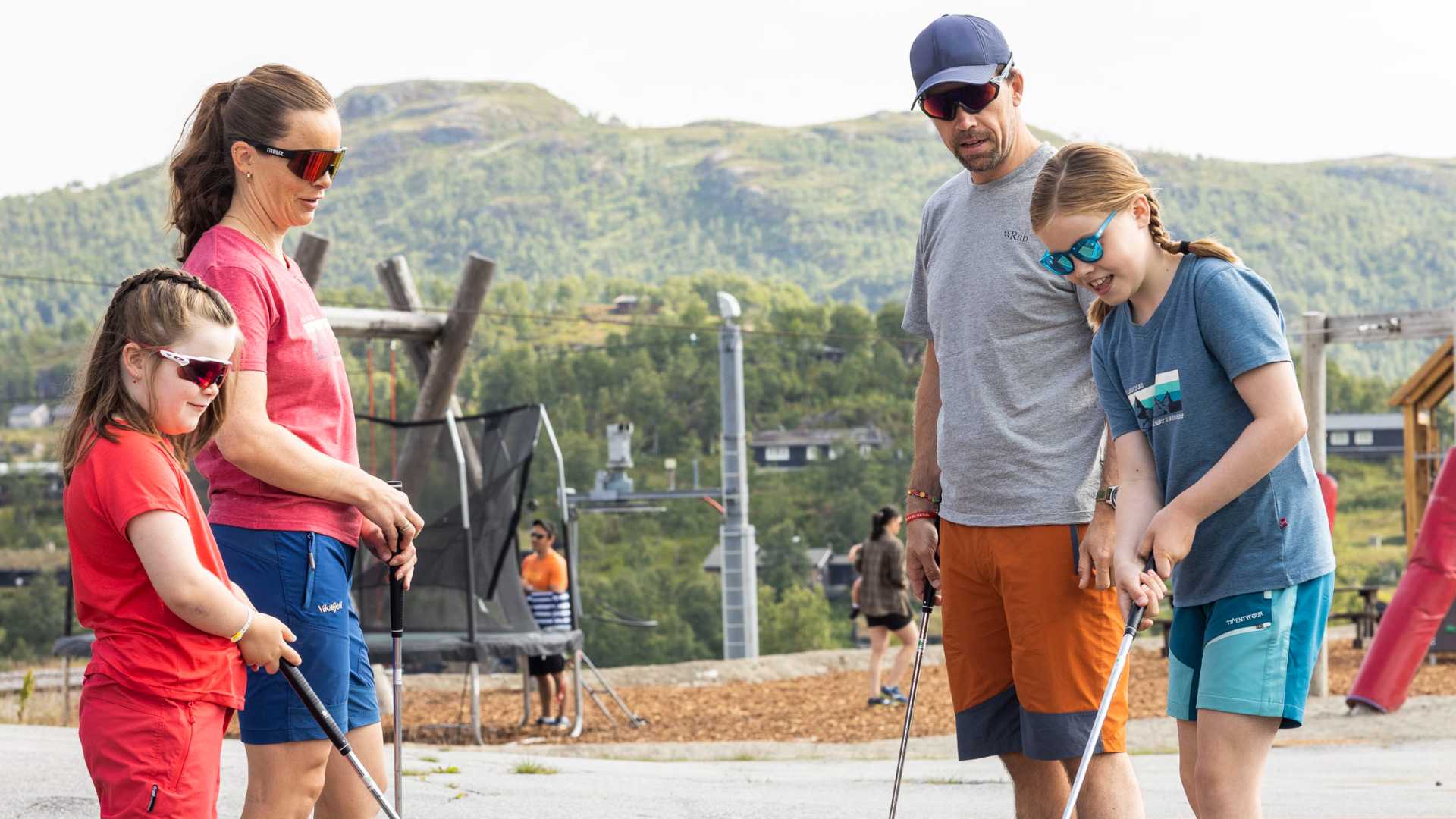 Familie nyter minigolf i fjellandskap, iført solbriller og fargerike klær, sommeraktivitet utendørs.