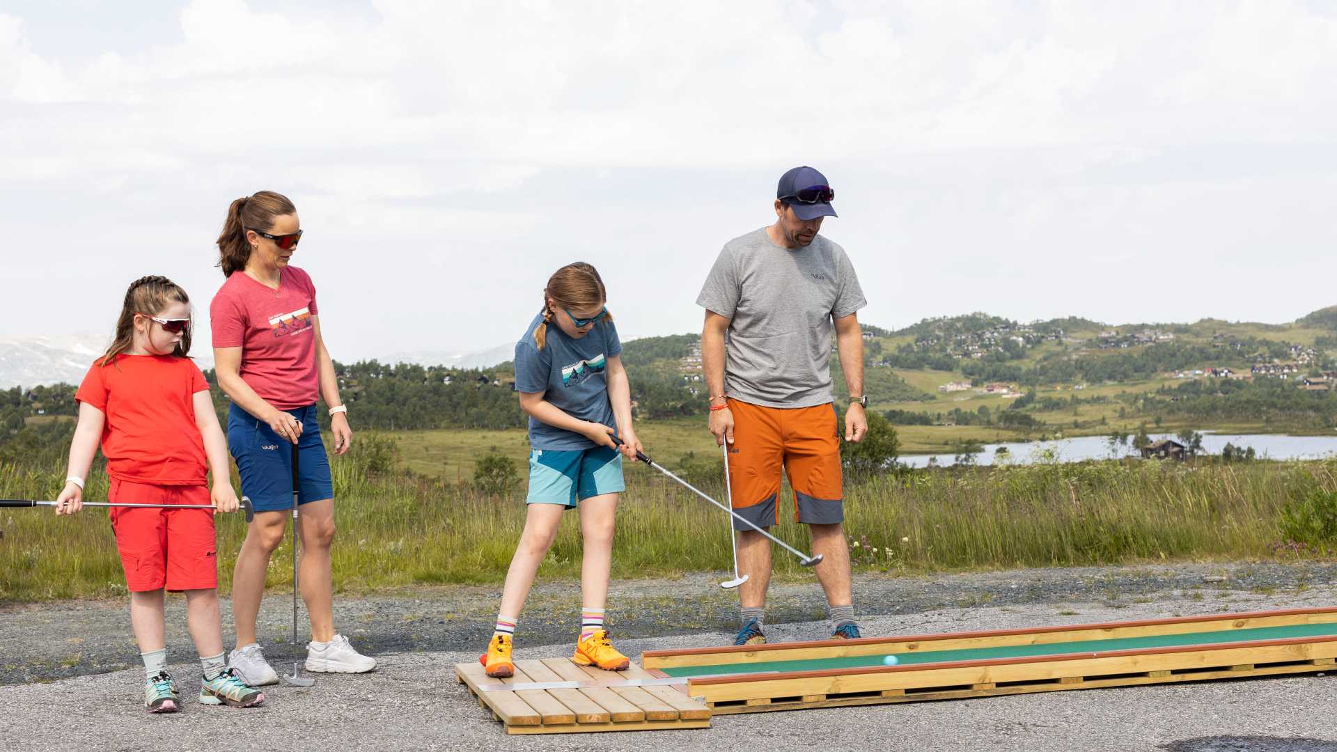 Familie spiller minigolf utendørs i solfylt norsk landskap, med fjell og innsjøer i bakgrunnen.