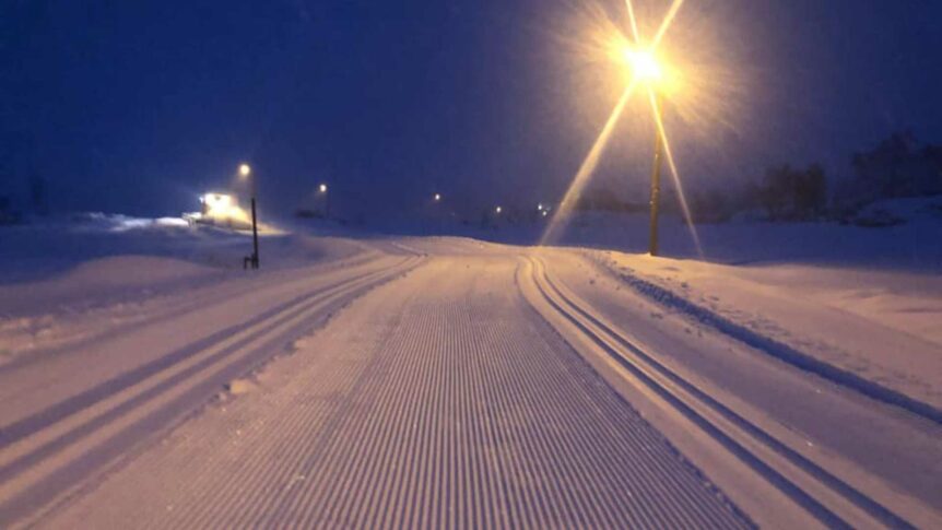 Nattbilde av opplyst langrennsløype i vinterlandskap, med lysmast og preparert snø.
