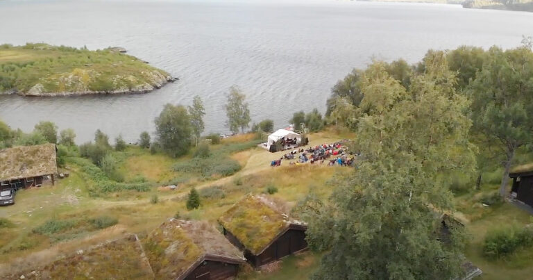Konsert ved innsjøen, folk samles ved scenen i naturskjønne omgivelser. Grønt landskap og gamle trehus i Norge.