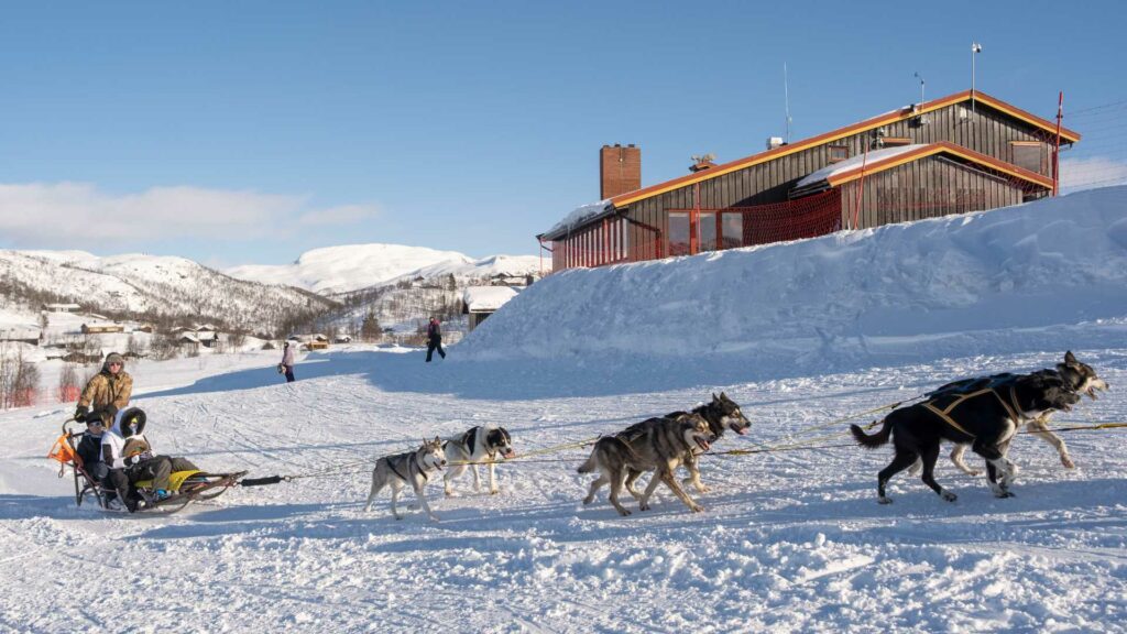 Hundeslede med mennesker i snødekte fjell, klar himmel i bakgrunnen.
