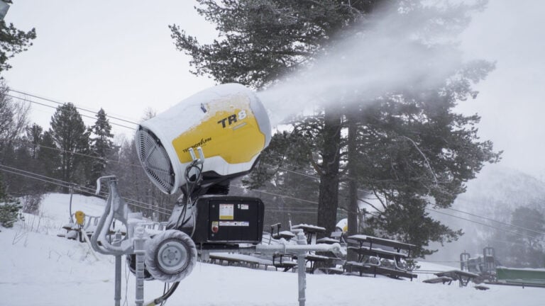 Snøkanon i drift på snødekt skianlegg, omgitt av furutrær og vinterlandskap.