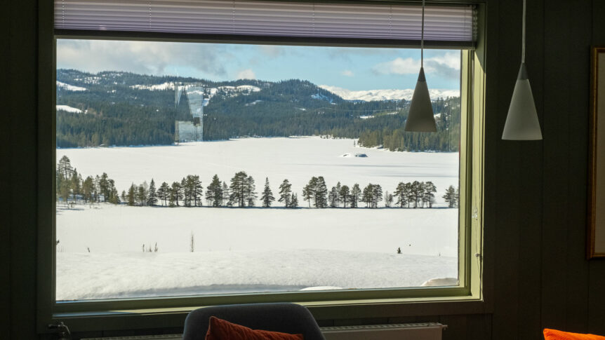 Utsikt mot snødekket landskap og trær gjennom stort vindu fra stue, vinterdag i naturen.