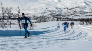 Langrennsløpere i konkurranse på snødekte fjell, med dramatiske vinterlandskap i bakgrunnen.