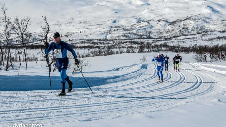 Langrennsløpere i konkurranse på snødekte fjell, med dramatiske vinterlandskap i bakgrunnen.
