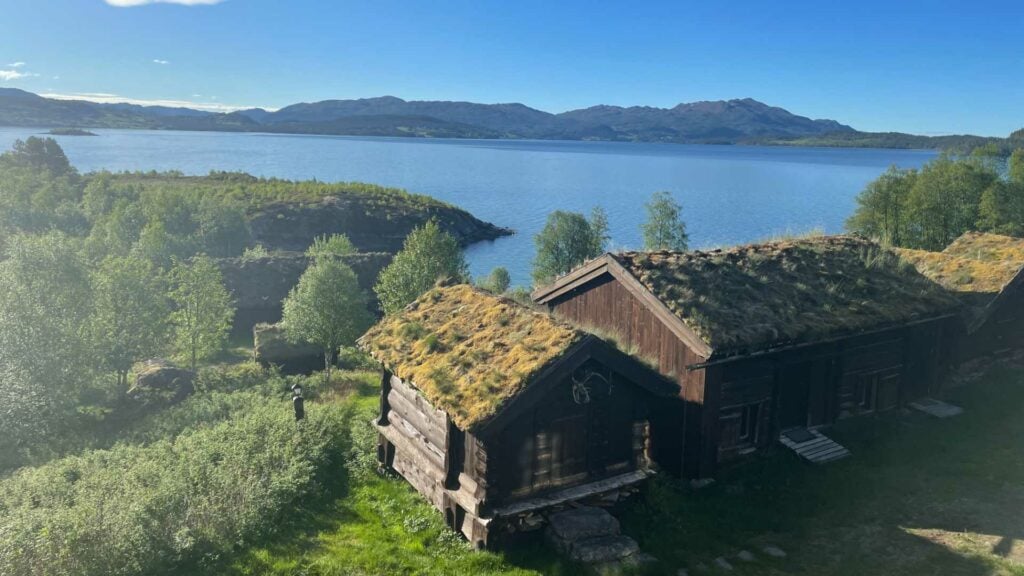 Gressdekkede hytter ved en innsjø med fjell i bakgrunnen og frodig natur på en solfylt dag.