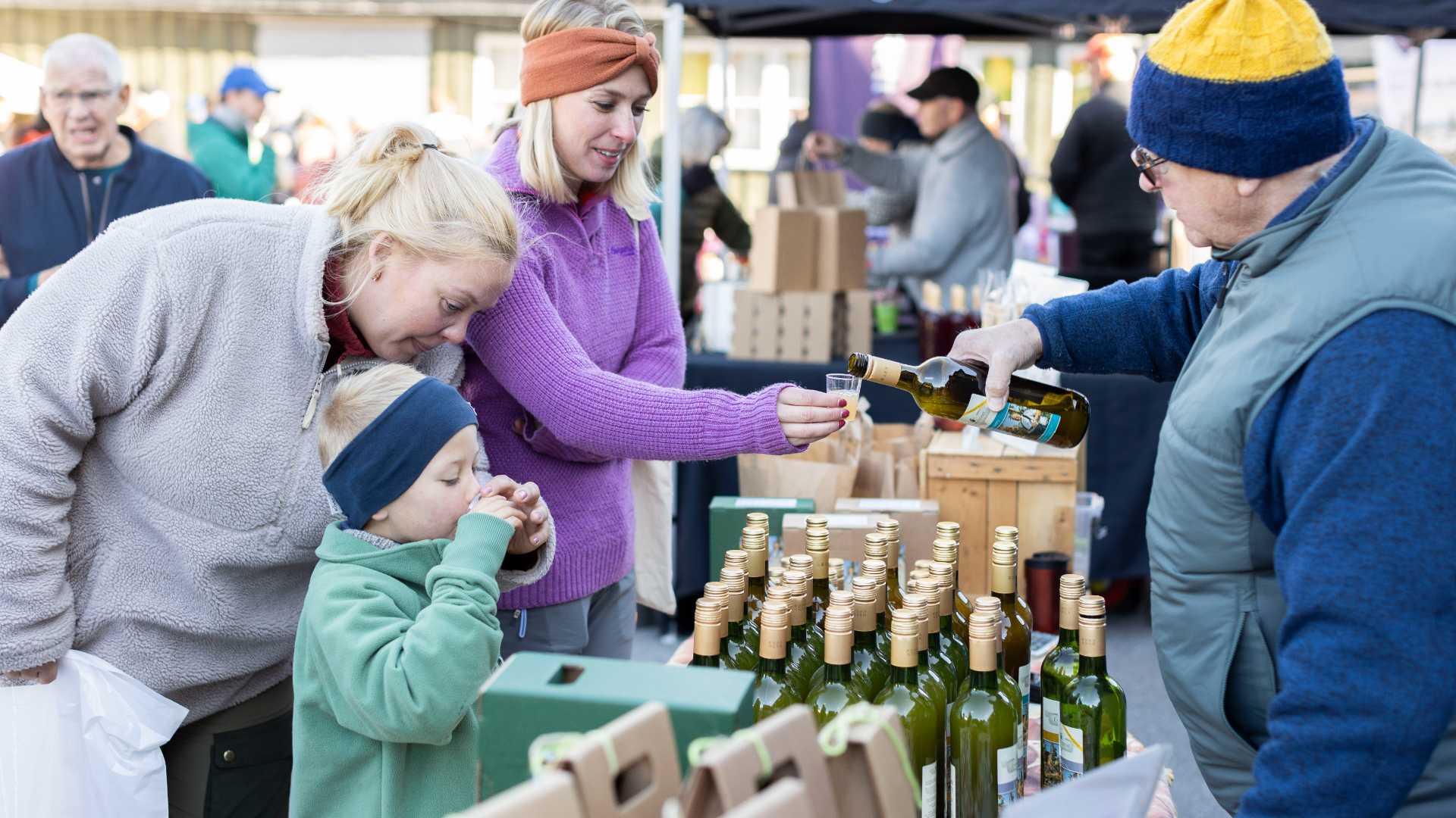 Familie smaker olivenolje på utendørs marked, flasker på bordet.