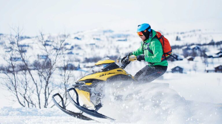 Mann i grønn jakke kjører snøscooter i snødekt landskap med fjell og trær i bakgrunnen.
