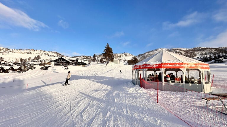 Snødekket skiområde med skiløper og paviljong på Rauland Skisenter, solfylt dag i fjellandskap.