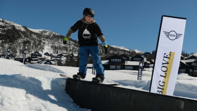 Barn snowboarder på rail i snødekkede fjell. Klar himmel, skiaktiviteter, vinterglede.
