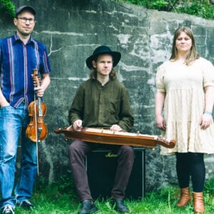 Tre musikere med instrumenter står og sitter ute mot en steinvegg, omringet av grønt gress og planter.