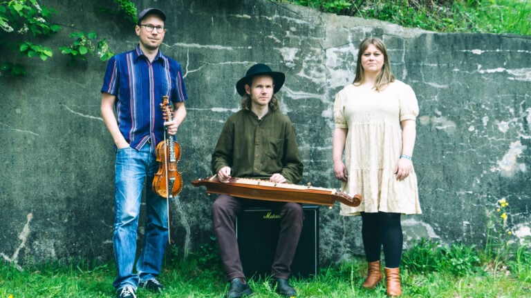 Tre musikere med instrumenter står og sitter ute mot en steinvegg, omringet av grønt gress og planter.