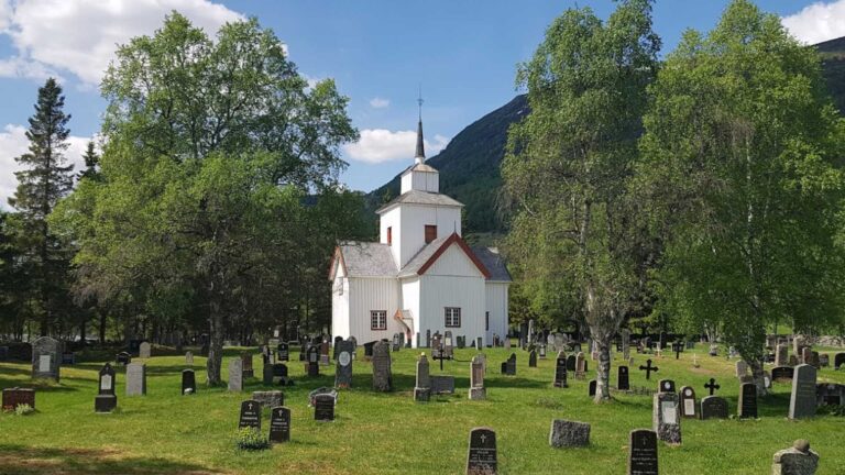 Hvit kirke omgitt av gravstøtter og trær i et grønt landskap under en klar blå himmel.