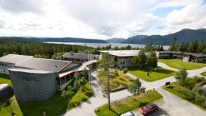 Utsikt over høyskolecampus ved fjord og fjellandskap i Norge, omgitt av skog under overskyet himmel.