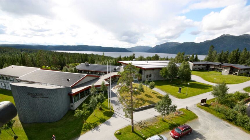 Utsikt over høyskolecampus ved fjord og fjellandskap i Norge, omgitt av skog under overskyet himmel.