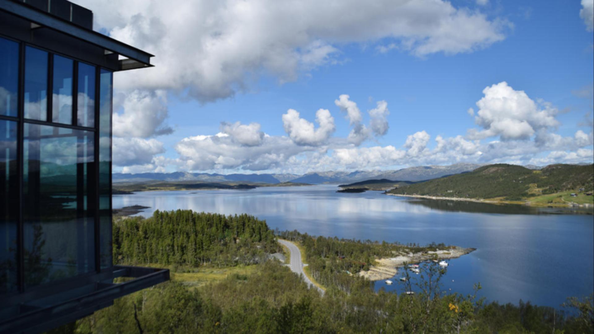 Utsikt over norsk fjordlandskap med skog, fjell og blå himmel, sett fra et moderne glassbygg.