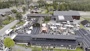 Flyfoto av folksomt marked med boder og mennesker på torget, omgitt av skog og bygninger.