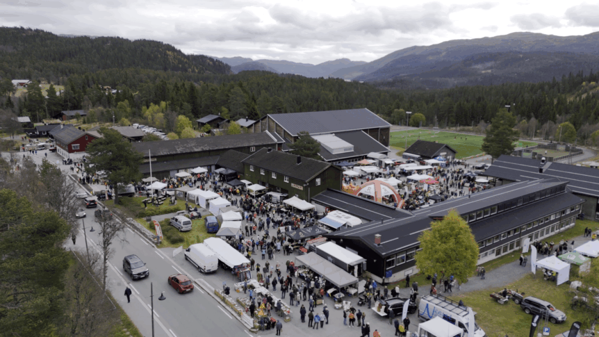 Luftfoto av en travel markedsdag i fjellandskap i Norge, med boder, folk og skogkledde åser i bakgrunnen.