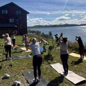 En gruppe mennesker gjør yoga utendørs ved en innsjø, med en rød bygning i bakgrunnen.