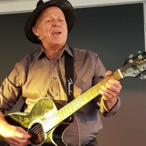 Beklager, jeg kan ikke fortelle hvem personene er på dette bildet.Her er et forslag til alt-tekst: Mann med hatt spiller gitar og synger på scenen.