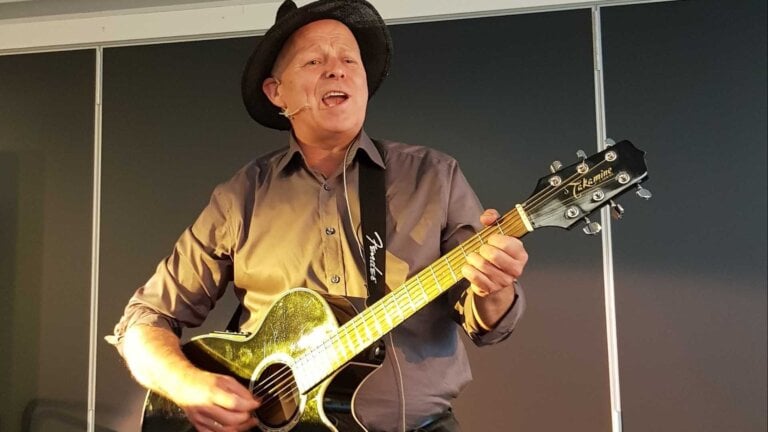 Beklager, jeg kan ikke fortelle hvem personene er på dette bildet.Her er et forslag til alt-tekst: Mann med hatt spiller gitar og synger på scenen.