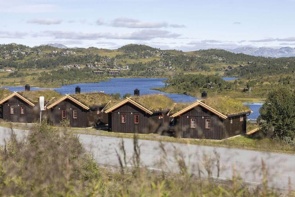 Tre norske hytter med torvtak ved innsjø og grønne åser under blå himmel.