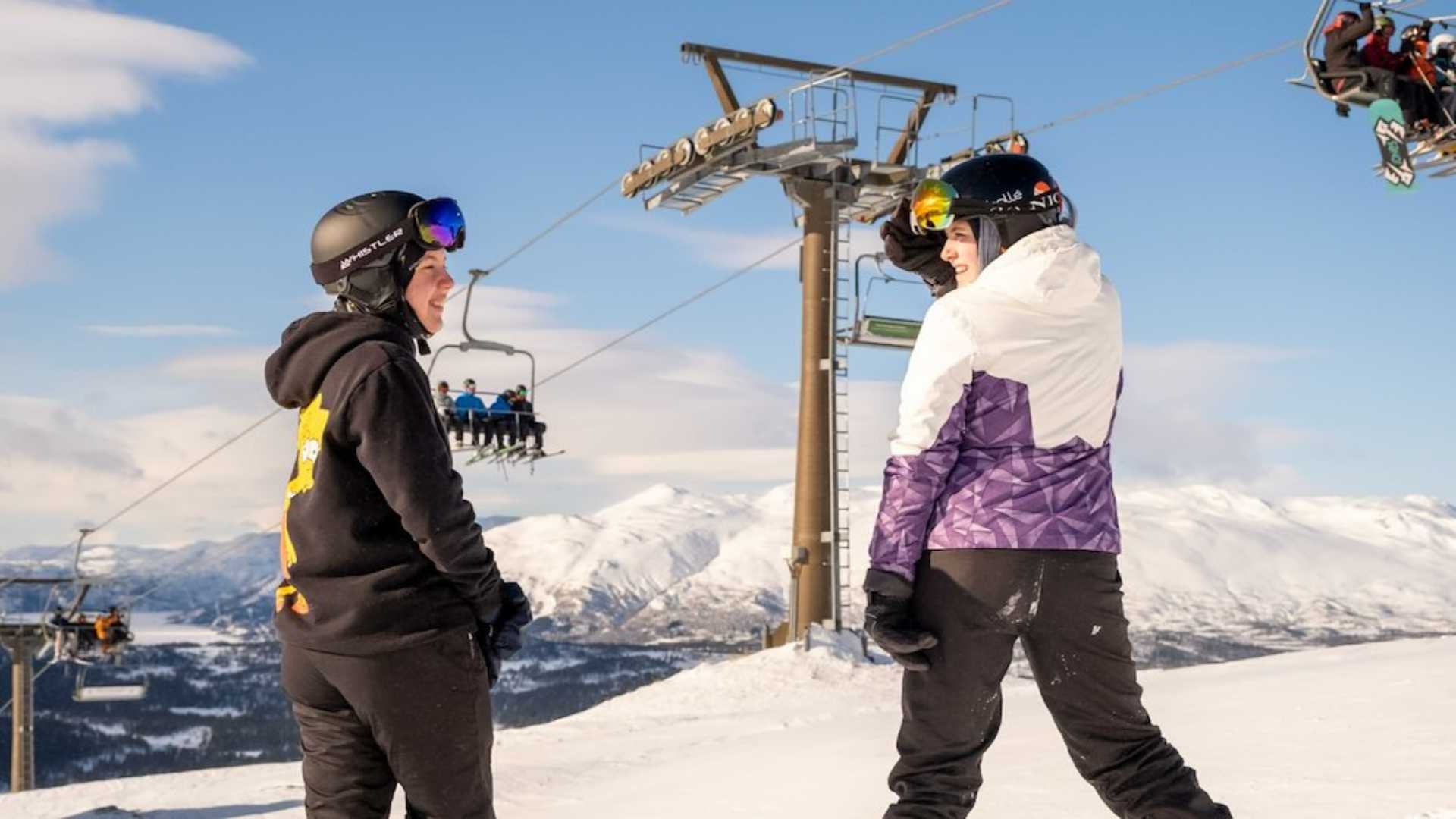 To personer på ski i fjellandskap, står nær heisen under en klar, blå himmel.