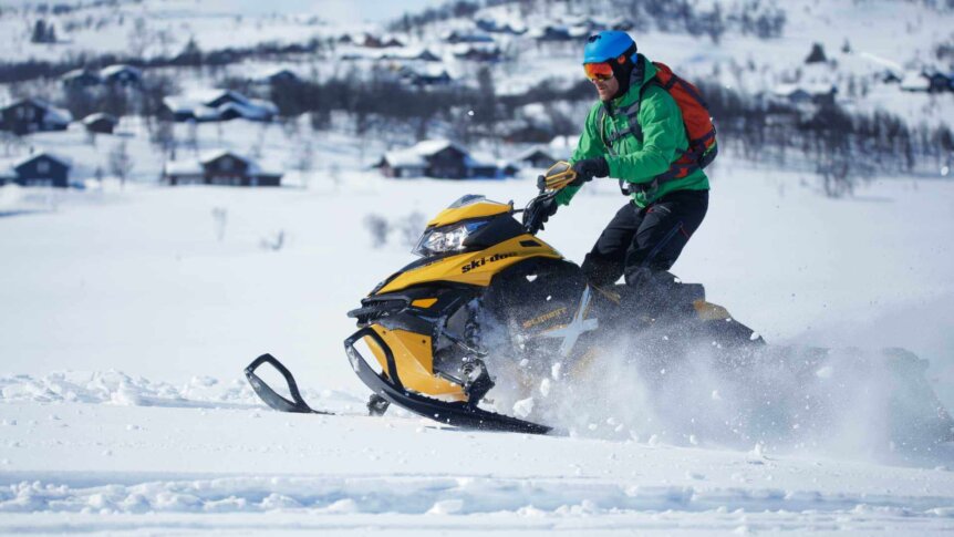 Mann kjører snøscooter i vinterlandskap med hytter i bakgrunnen, iført grønn jakke og blå hjelm.