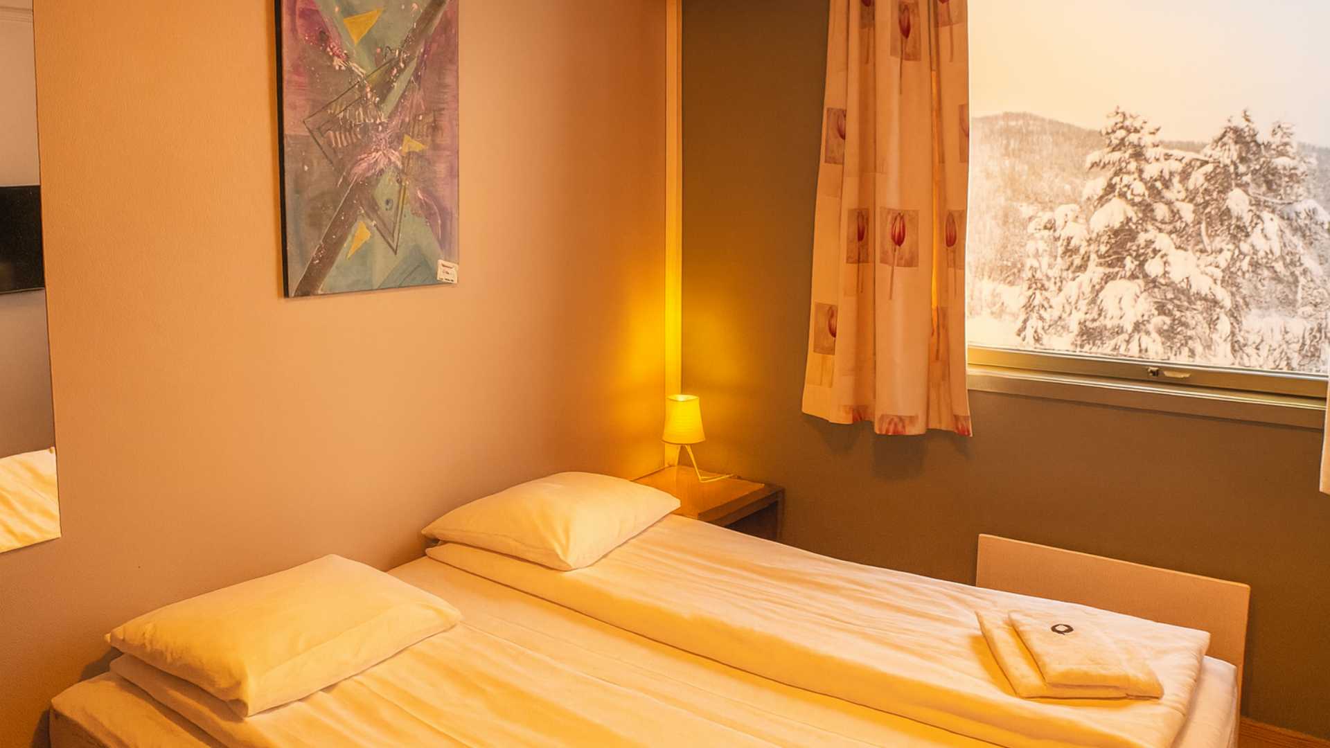 Koselig hotellrom med seng, natty lampe og vinterutsikt gjennom vinduet til snødekte trær og fjell.