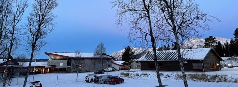 Vinterlandskap med snødekte bygninger, trær og fjell i bakgrunnen under den blå himmelen.