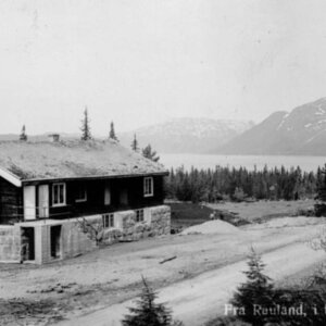 Gammelt ungdomshus i fjellandskap, med trær og fjord i bakgrunnen i Retland, Norge. Svart-hvitt bilde.