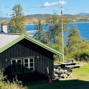 Svart hytte ved sjøen med norsk flagg og piknikbord, omgitt av natur og blå himmel.