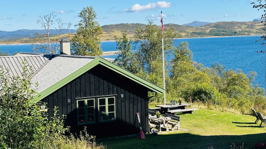 Svart hytte ved sjøen med norsk flagg og piknikbord, omgitt av natur og blå himmel.