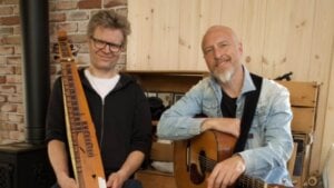 To menn med musikkinstrumenter, en har en gitar og den andre en langeleik. Brick vegg og trepanel i bakgrunnen.